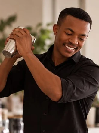 A smiling bartender in a black shirt shakes a cocktail shaker at shoulder height.